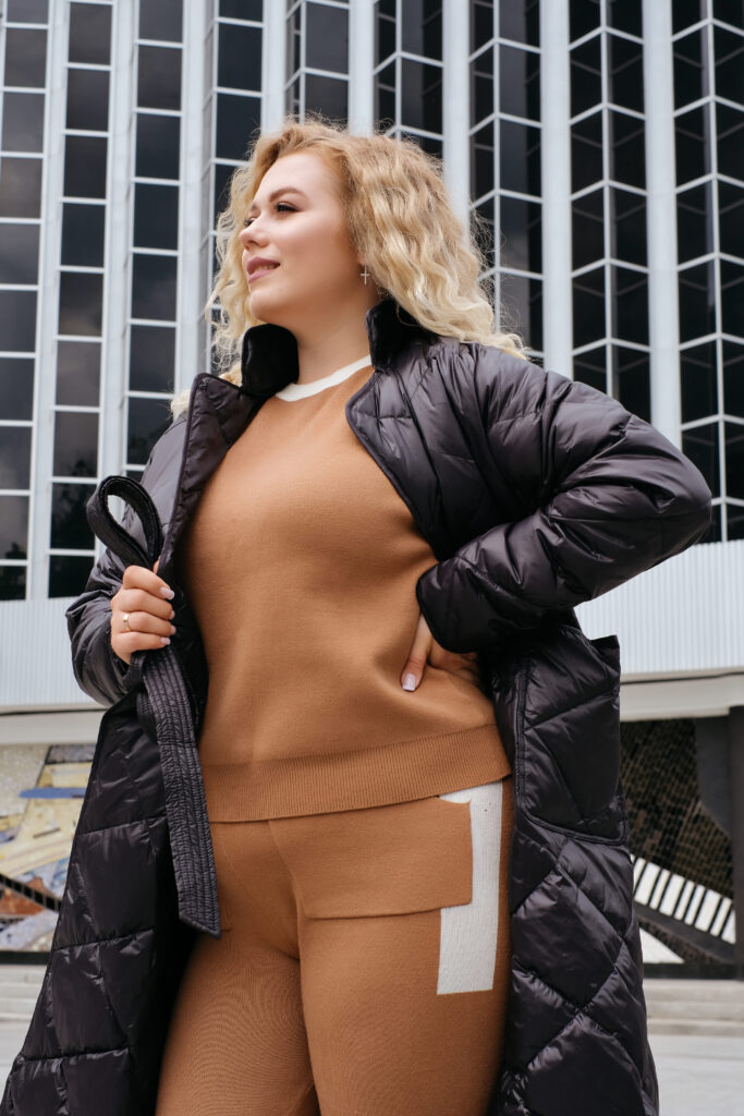 Blonde plus-size model in a trendy black oversized quilted coat, posing during a city walk for an advertising banner