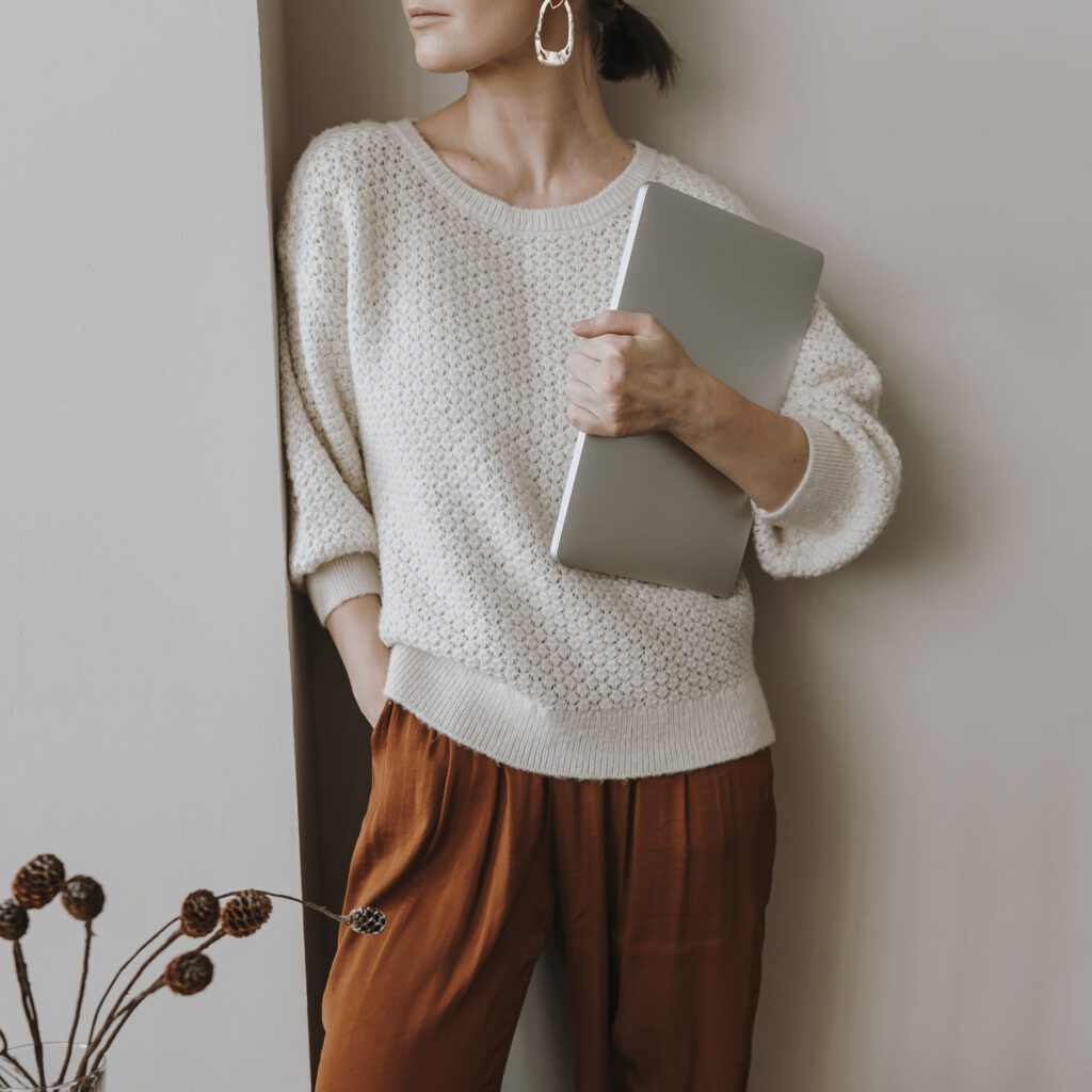 Woman dressed in a neutral-colored sweater and earth-toned pants, holding a laptop