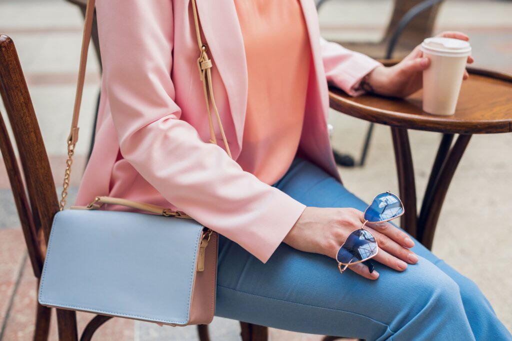 Close-up of a stylish woman's accessories as she sits in a café, featuring sunglasses, a handbag, and pink and blue hues, embodying spring-summer fashion trends and elegance while enjoying a coffee in her work attire.