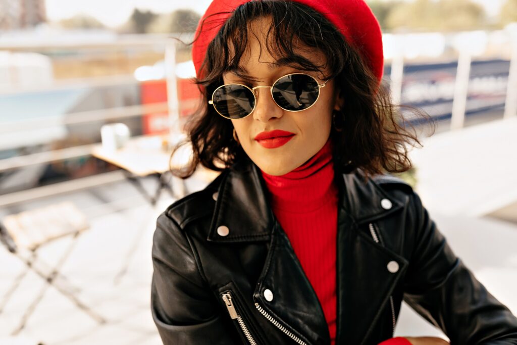 Close-up image of an endearing, charming woman with red lipstick and sunglasses, beaming as she waits for her friends at an outdoor cafe.
