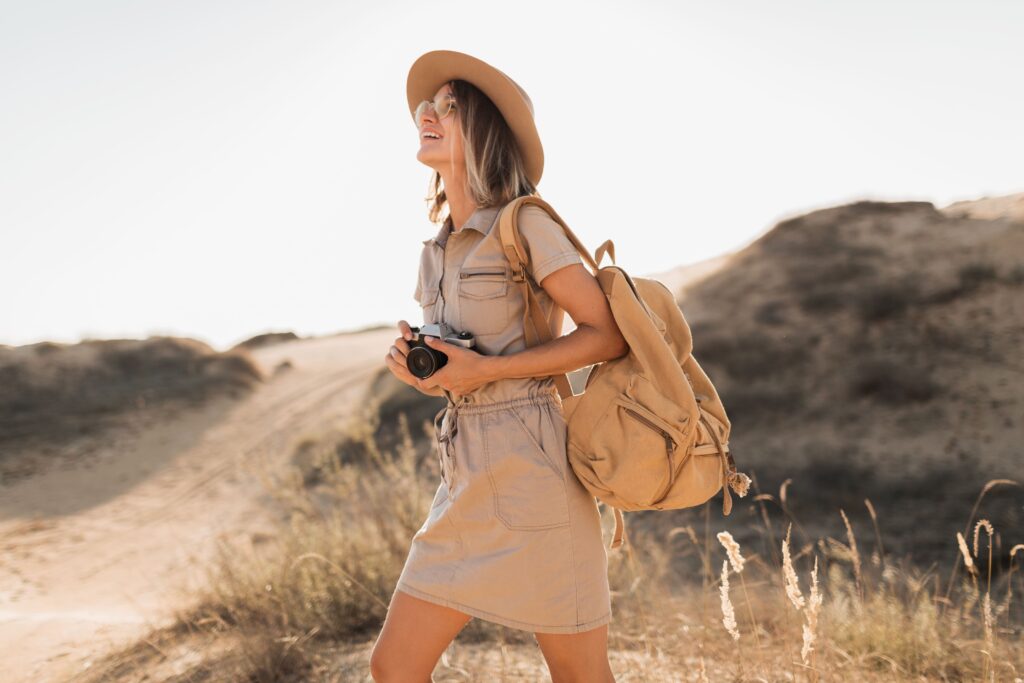 Attractive stylish young woman in khaki dress in desert, traveling in africa on safari, wearing hat and backpack, taking photo on vintage camera