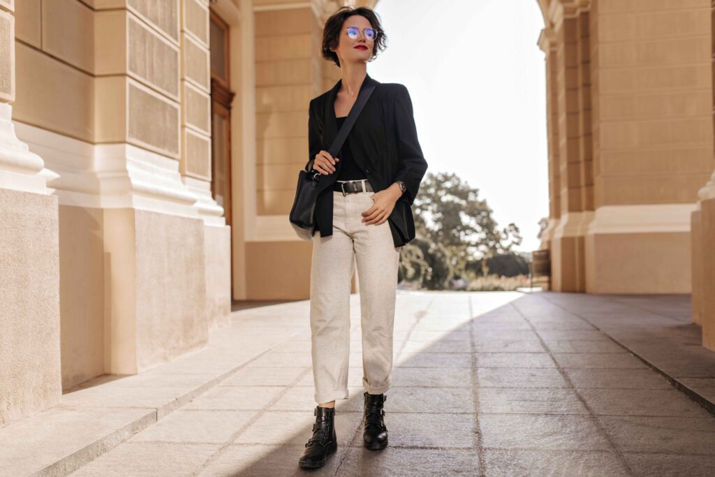 Full-length image of a woman in a minimalist outfit, composed of light trousers, boots, a jacket, black handbag, and eyeglasses, smiling on a street.