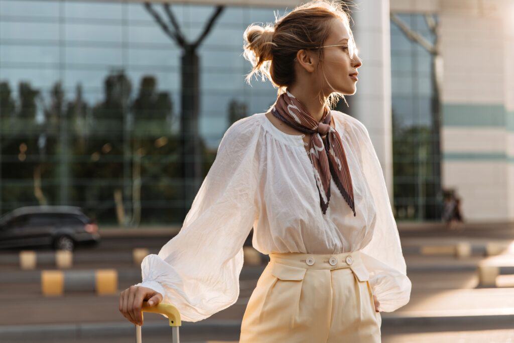 A portrait of a blonde girl wearing a white blouse, beige pants, a brown silk scarf, and eyeglasses. She is holding her luggage and gazing into the distance.