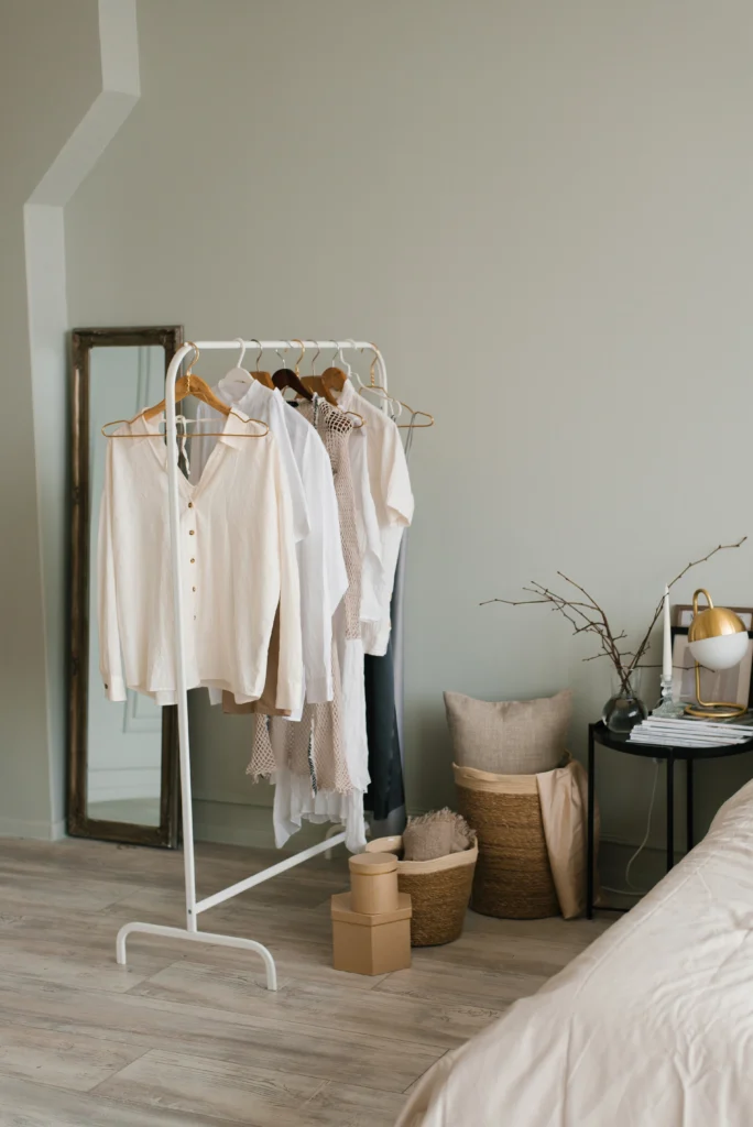 A modern, open dressing room featuring essential elements of a minimalist wardrobe. It's neatly organized with a selection of women's clothing hung up on a rack, showcasing staple pieces like a crisp white shirt, black trousers, and a simple dress. Below, a shelf neatly displays a range of stylish shoes. The image epitomizes the concept of a clean, versatile, and timeless minimalist wardrobe.