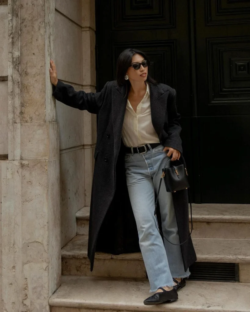 Minimalist woman wearing a brown trench coat with silk white blouse and denim jeans with a hand bad and sunglasses