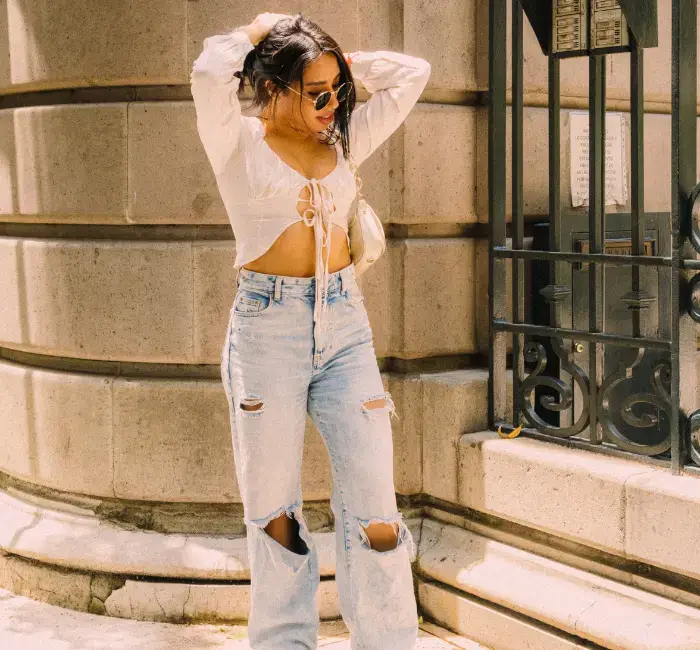 Stylish woman in a white crop top and distressed jeans, accessorized with sunglasses and a crossbody bag, showcasing a trendy and casual outfit.