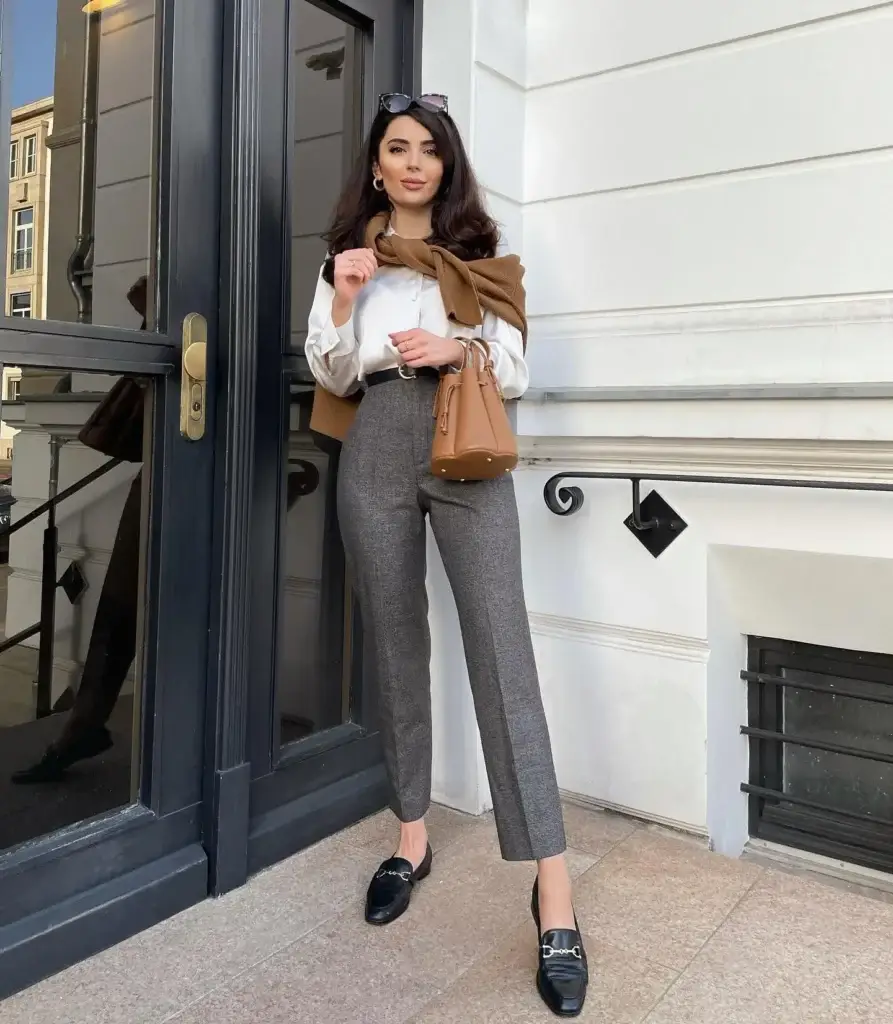 "A woman poses elegantly on a city sidewalk, exemplifying Parisian (French-Girl) style. She wears a crisp white blouse and tailored grey trousers, accented with a camel-colored scarf draped over her shoulders. Her accessories include a matching camel handbag, sleek black loafers, and sunglasses perched atop her head. The background features classic architecture with large black doors and white walls, enhancing the sophisticated urban setting of the photograph.