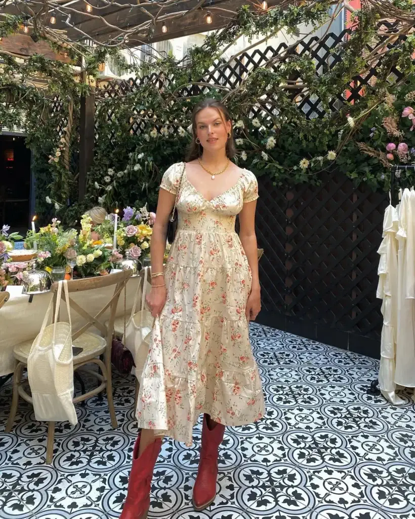 A woman stands in a whimsical garden setting, wearing a romantic cottagecore-style dress adorned with a delicate floral print. The dress features a tiered skirt and puff sleeves, paired with striking red boots. Her look is completed with a simple necklace and earrings, perfectly blending rustic charm and elegance.