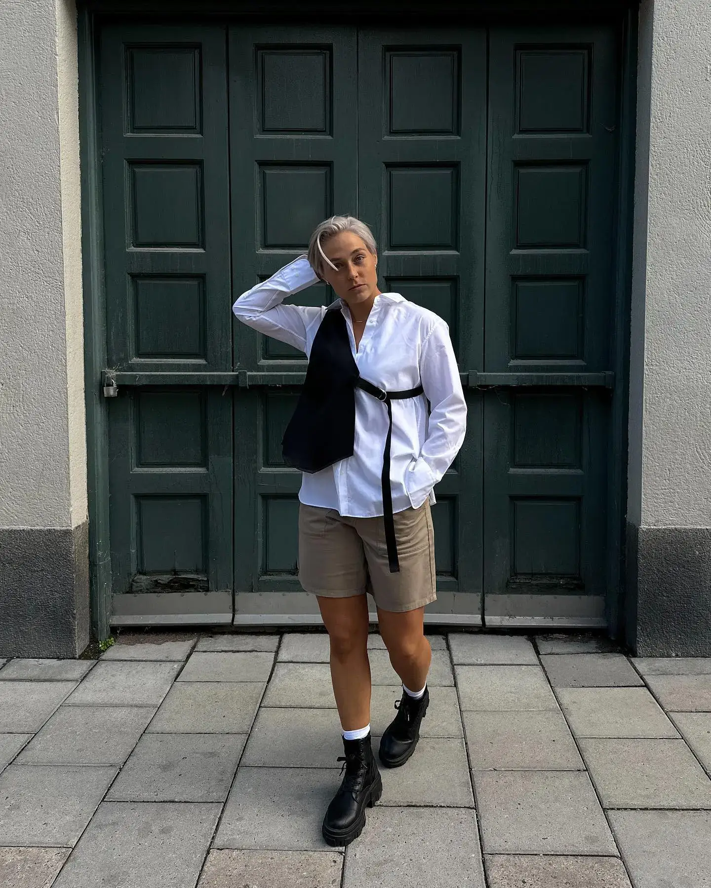 A person stands confidently against a dark green door, showcasing an androgynous style. They wear a crisp white button-up shirt under a black sleeveless vest, paired with beige tailored shorts. The outfit is completed with chunky black combat boots and white socks. A minimalistic approach and neutral colors emphasize the sleek, gender-neutral fashion look.