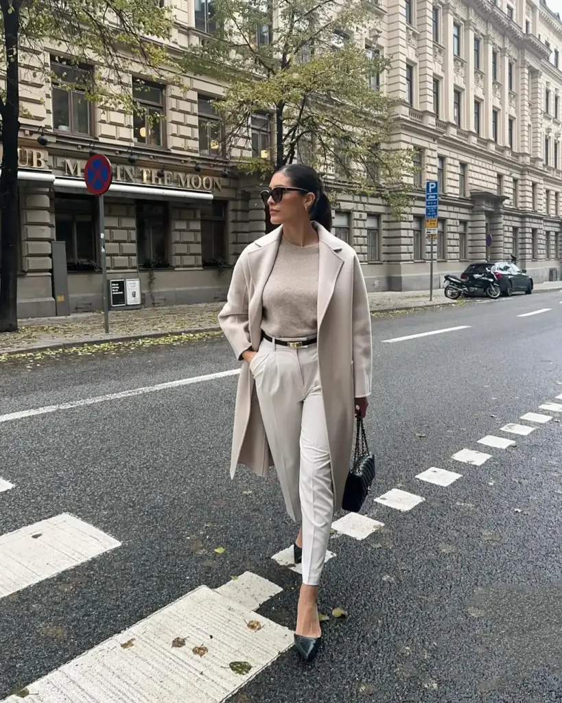 A stylish woman in a classic autumn outfit walks confidently across a street in an urban setting. She wears a light grey oversized coat over a beige turtleneck sweater, paired with high-waisted cream trousers and a black belt. Her accessories include black pointy-toe heels, dark sunglasses, and a quilted black shoulder bag. Behind her, the city street features a historical building with intricate architecture, a 'no parking' sign, and sparse fall foliage on trees, adding to the autumnal ambiance.