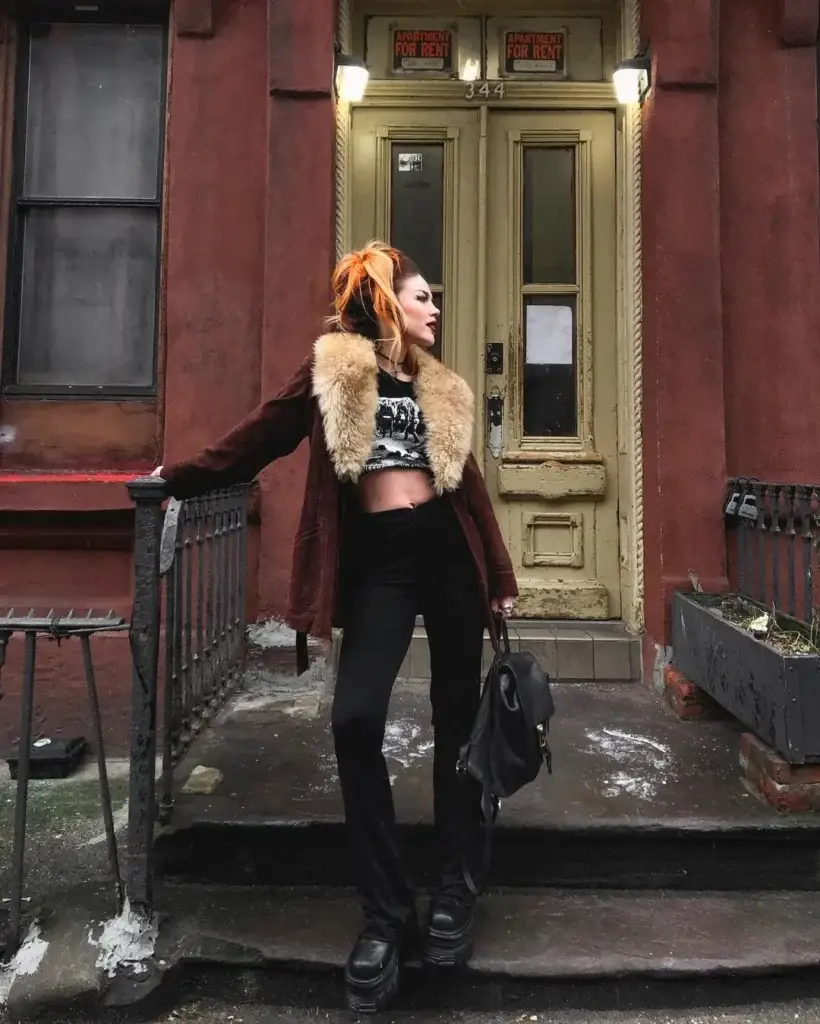 A woman poses confidently on a city street, sporting a grunge-inspired outfit. She wears a cropped graphic tee under a fur-lined burgundy jacket, paired with black high-waisted pants and chunky black platform boots. Her orange hair is styled up, and she carries a black backpack, standing in front of a brownstone building with 'Apartment for Rent' signs in the background.