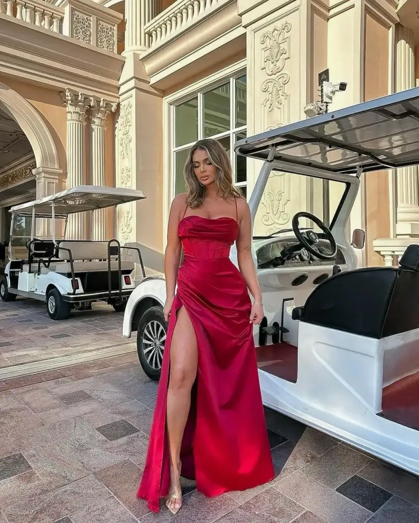 A woman in a romantic style poses elegantly in front of an ornate building with a golf cart nearby. She wears a stunning red satin gown with a sweetheart neckline and a high slit, showcasing her toned legs. The dress is complemented by delicate strappy heels, adding a touch of sophistication to her glamorous look.