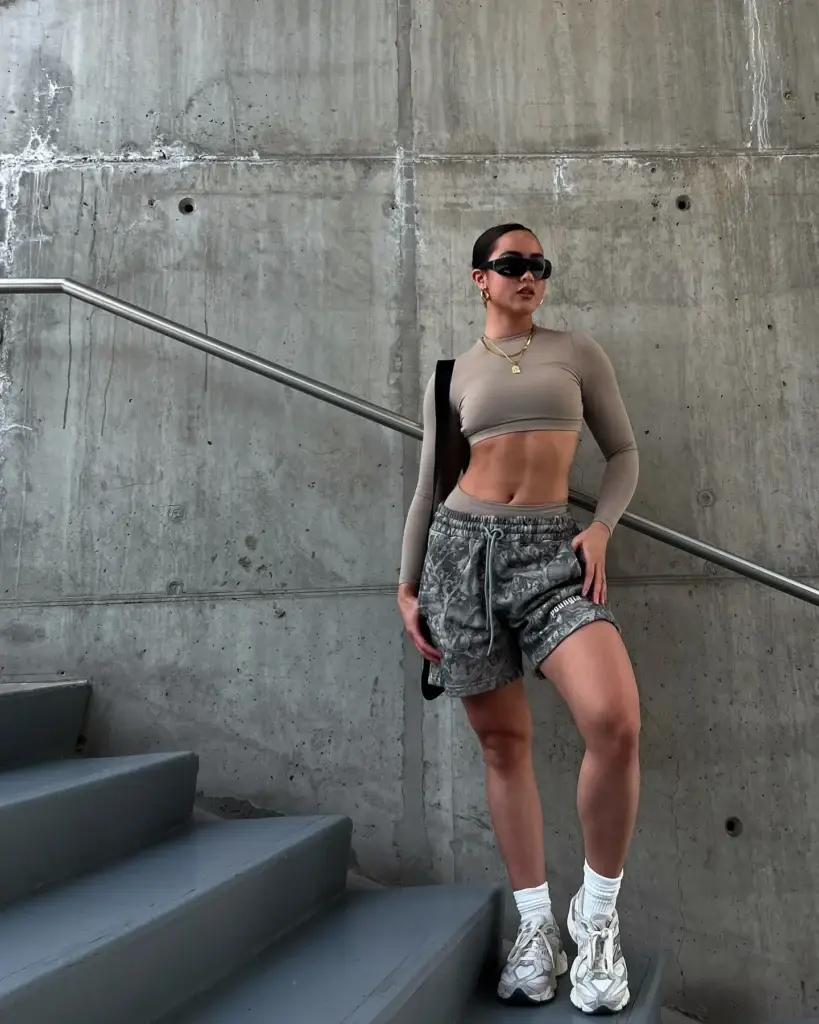 A young woman stands confidently on a concrete staircase, showcasing her athleisure style. She wears a form-fitting taupe crop top paired with high-waisted camo print shorts and chunky white sneakers. Accessories include a black shoulder bag, gold necklace, and stylish black sunglasses, complementing her sporty yet chic look.