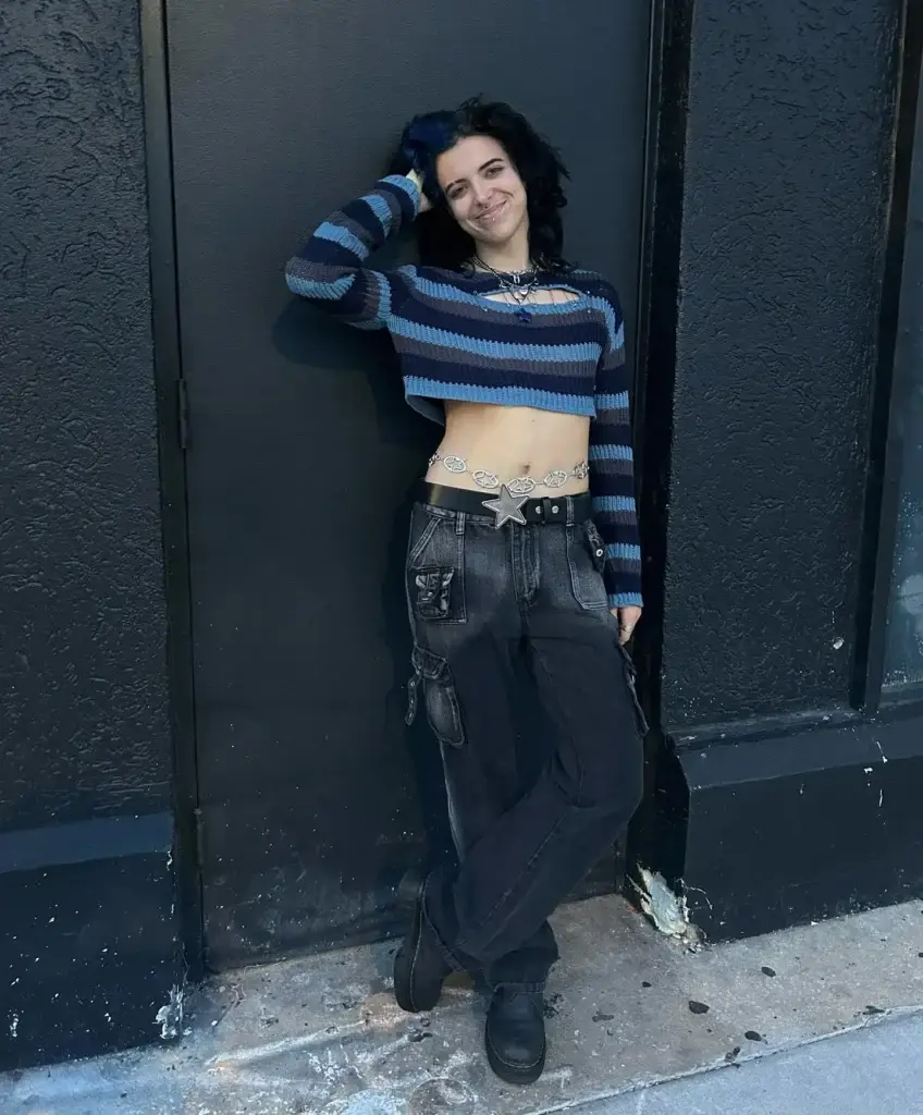 A woman leans against a dark textured wall, showcasing her punk rock style. She sports a cropped, long-sleeve striped sweater in blue and black, paired with distressed black jeans featuring large pockets and chain details. Her look is completed with chunky black boots, a studded belt, and vibrant blue hair, echoing a rebellious yet playful vibe.