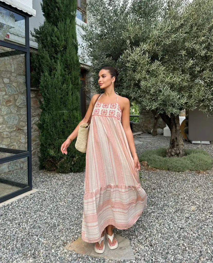 A woman stands in a serene garden, embodying eco-friendly style in a flowing maxi dress. The dress features soft pink and white stripes with intricate red embroidery around the neckline. She accessorizes with a simple straw tote bag and white sandals, complementing her natural, sustainable fashion look.