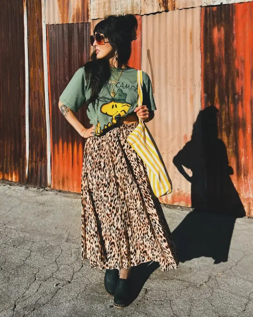 A woman stands confidently against a rustic metal backdrop, sporting a vibrant outfit with a green graphic tee tucked into a leopard print skirt. She complements her bold look with yellow-striped accessories, clog boots, and stylish sunglasses, exuding a chic vintage vibe.