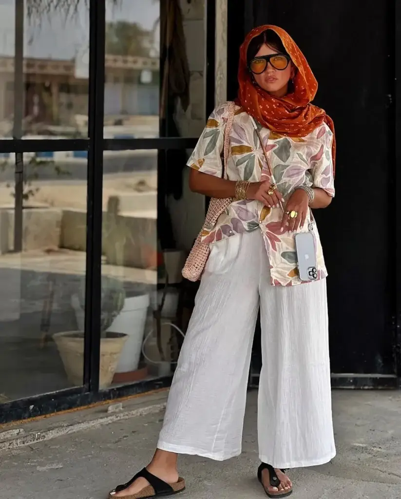 A woman wearing a Bohemian-style outfit, standing in front of a glass door. She is dressed in white, wide-legged pants and a patterned blouse with a dominant leaf design, complemented by an orange headscarf and large sunglasses. Her look is accessorized with multiple bangles, a woven bag, and flat sandals.