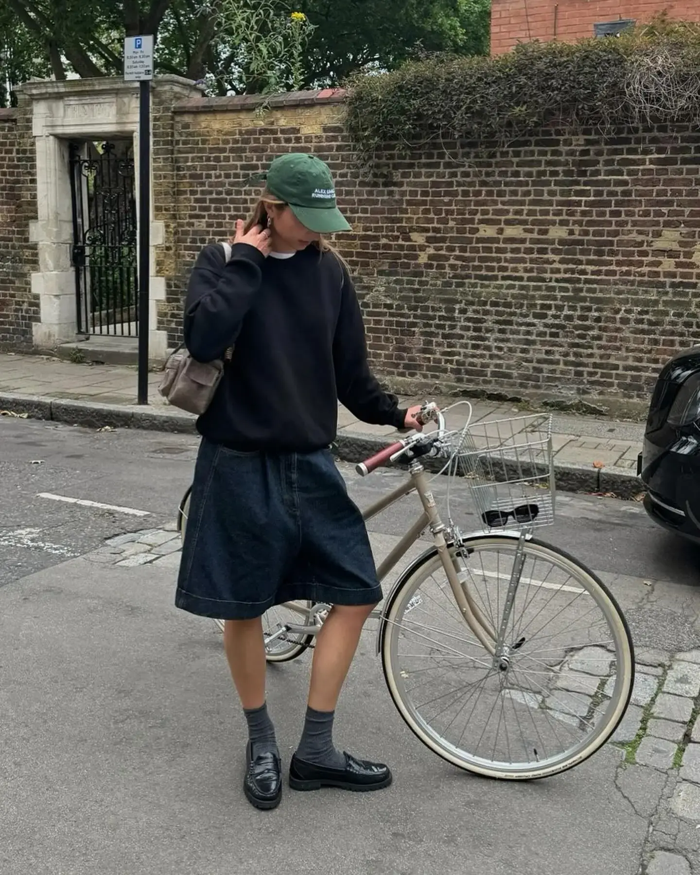 A young person in a relaxed street style outfit, wearing a black sweatshirt, dark denim knee-length shorts, and black loafers paired with grey socks. They are adjusting a green baseball cap and holding onto a vintage bike, standing on a cobblestone street beside a brick wall. Their casual attire is complemented by a practical shoulder bag, exuding a cool, urban vibe.