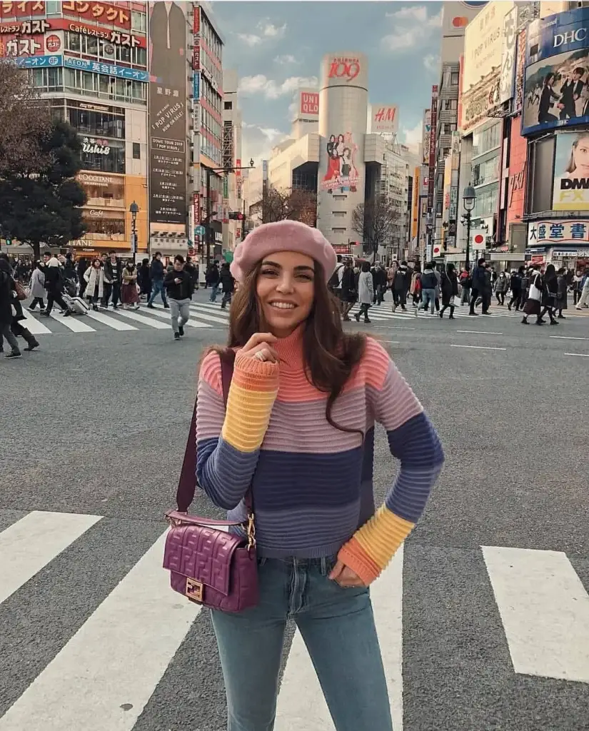 A young woman in a vibrant, youthful outfit stands on a busy street in Tokyo, Japan. She wears a pastel-striped, long-sleeve top with a pink beret, exuding a playful and chic gamine style. She accessorizes with a purple crossbody bag and smiles warmly at the camera, with bustling city life and colorful advertisements in the background.