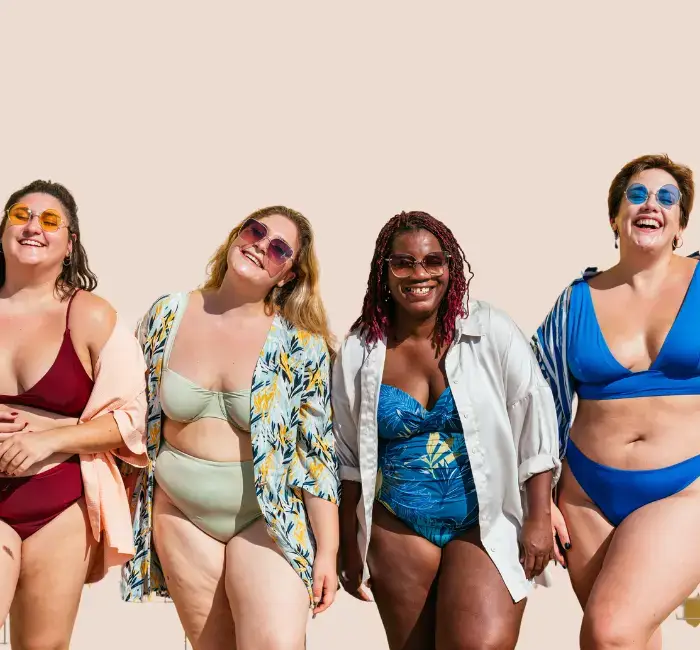 Four joyful women of diverse curvy body types posing in stylish swimwear against a pale backdrop, each wearing sunglasses and showcasing a range of vibrant, body-positive beach attire.