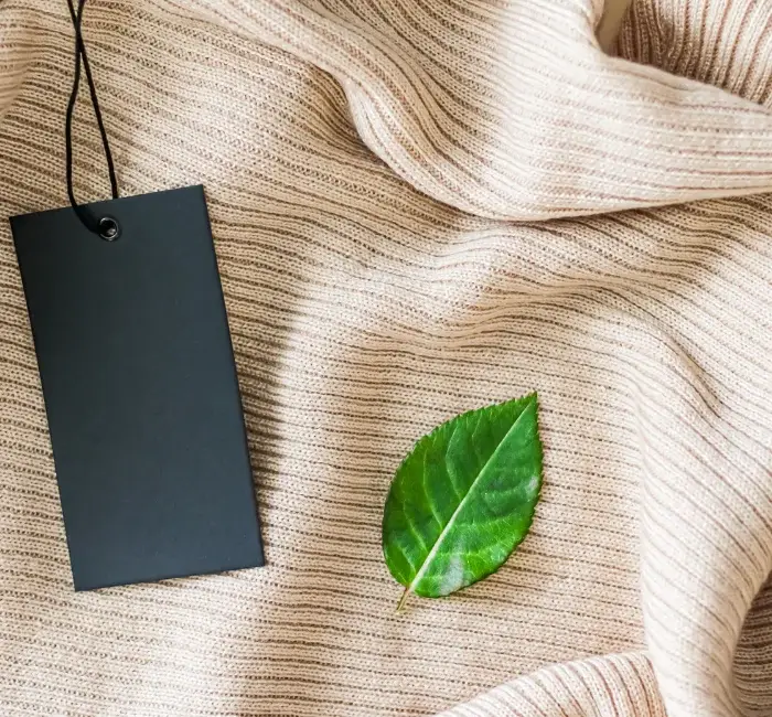 A black clothing tag and a green leaf rest on a beige knitted fabric, symbolizing sustainable fashion and eco-friendly practices.