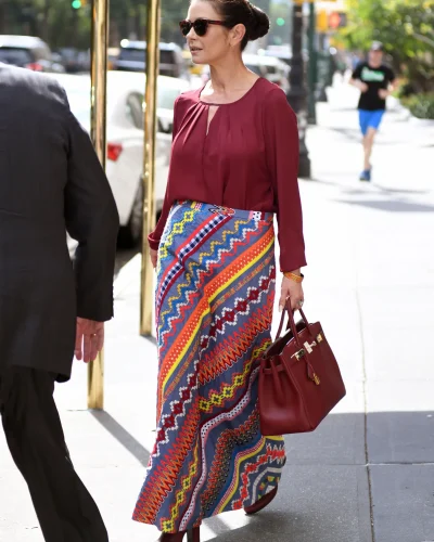 Catherine Zeta-Jones, a celebrity with an apple-shaped body, wearing a burgundy long-sleeved blouse tucked into a colorful, patterned maxi skirt. She accessorizes with a matching burgundy handbag and sunglasses. Her hair is styled in a sleek bun, and she walks confidently on the sidewalk, showcasing her chic and vibrant fashion sense.