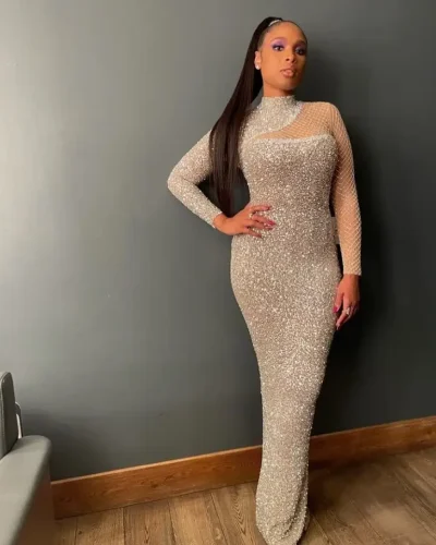 A stylish woman with an apple-shaped body wearing a sparkling, long-sleeved, form-fitting silver gown with mesh detailing around the neckline and sleeves. She poses confidently with one hand on her hip, showcasing her elegant and glamorous look.