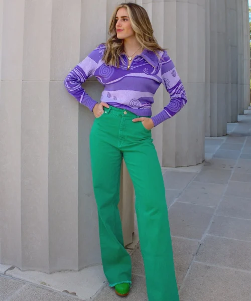 A woman wearing a purple sweater with a swirl pattern and green pants, standing confidently with her hands on her hips in front of a light gray, columned building.