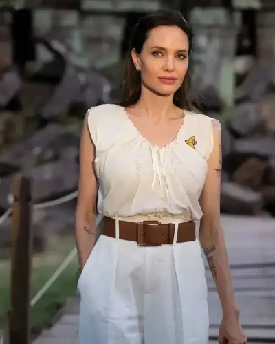 Angelina Jolie, a celebrity with an apple-shaped body, wearing a sleeveless white blouse with a small butterfly detail, tucked into high-waisted white pants with a wide brown belt. She stands confidently outdoors, with a serene expression, showcasing her chic and casual style. Her hair is styled back, and she has minimal makeup, emphasizing her natural beauty.