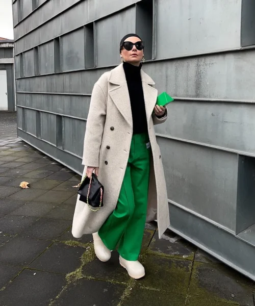 A woman wearing green wide-legged pants, a black turtleneck, and a beige overcoat. She accessorizes with black sunglasses, a small black handbag, and a green wallet, walking confidently against a modern industrial background.