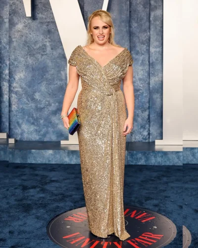 Rebel Wilson, a celebrity with an apple-shaped body, wearing a stunning gold sequin gown with a draped, off-the-shoulder neckline at the Vanity Fair event. She accessorizes with a colorful clutch and subtle jewelry. Her blonde hair is styled in a chic updo, and she exudes confidence and glamour against the sophisticated backdrop.