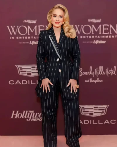 Adele, a celebrity with an apple-shaped body, wearing a stylish black pinstripe suit with a tailored blazer and wide-leg pants at the Women in Entertainment event. She accessorizes with bold gold earrings and rings, and her blonde hair is styled in glamorous waves. She stands confidently against a maroon backdrop with event logos, showcasing her chic and sophisticated fashion sense.