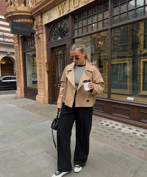 Woman wearing a casual chic dress code outfit with a beige trench coat, striped top, black wide-leg trousers, and black and white sneakers, holding a coffee cup and small black handbag, standing in front of a shop.