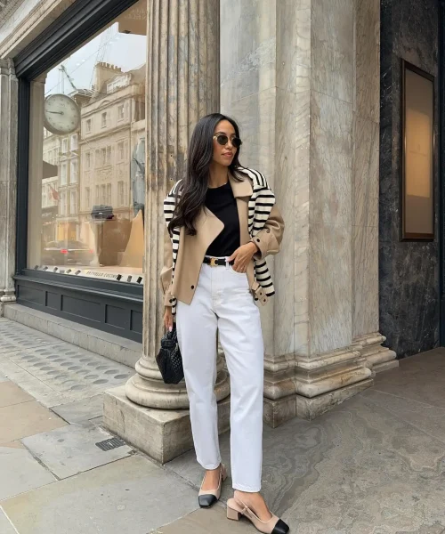 Woman wearing a casual chic dress code outfit with a black top, beige trench coat, white jeans, beige and black heels, and a black handbag, standing in front of a stone building.