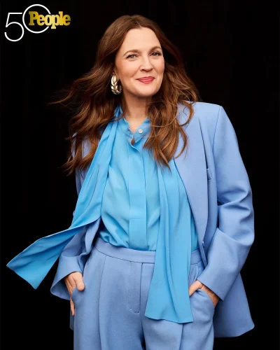 Drew Barrymore, a celebrity with an apple-shaped body, is dressed in a stylish blue pantsuit with a matching blouse and scarf. She has her hands in her pockets and wears large gold earrings. Her long, wavy hair cascades over her shoulders, and she smiles confidently. The image is from People magazine's "50 Most Beautiful People" feature.