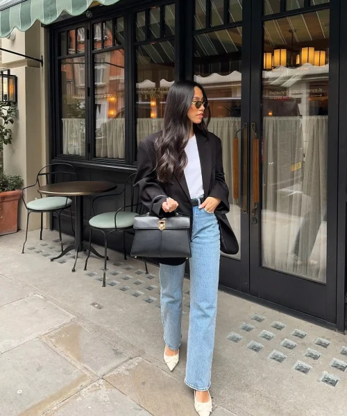 Woman wearing a casual chic dress code outfit with a white top, black blazer, light wash jeans, white heels, and a black handbag, walking past a cafe.