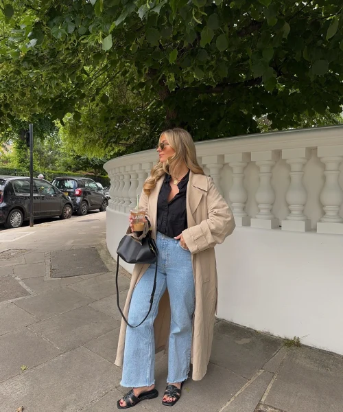 Woman wearing a casual chic dress code outfit with a black blouse, light wash wide-leg jeans, beige trench coat, and black sandals, holding a black handbag and a drink, standing on a sidewalk in front of a white fence and greenery.