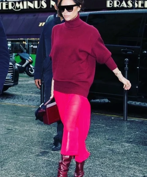 A stylish woman dressed in a maroon turtleneck sweater and vibrant red skirt, accessorized with maroon knee-high boots and dark sunglasses, walks confidently on a city street, carrying a maroon handbag.