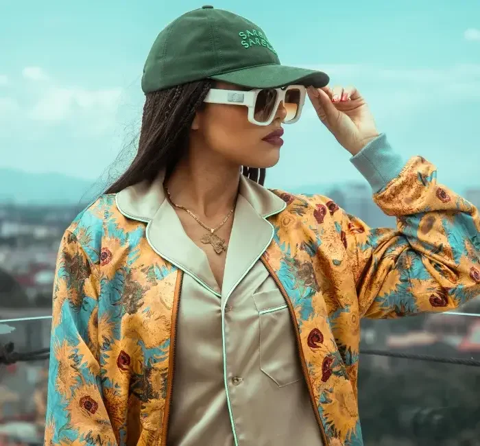 A stylish woman wearing a green baseball cap, oversized sunglasses, a satin shirt, and a colorful floral jacket poses outdoors against a cityscape backdrop, showcasing affordable sustainable clothing with a vibrant and trendy vibe.