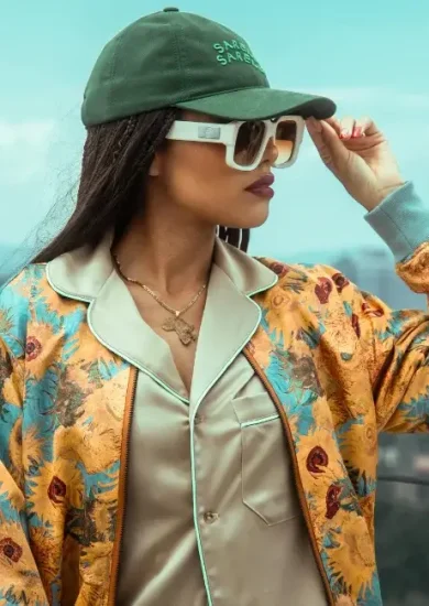 A stylish woman wearing a green baseball cap, oversized sunglasses, a satin shirt, and a colorful floral jacket poses outdoors against a cityscape backdrop, showcasing affordable sustainable clothing with a vibrant and trendy vibe.