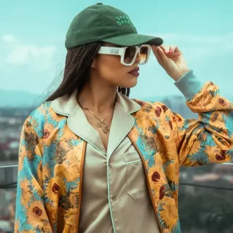 A stylish woman wearing a green baseball cap, oversized sunglasses, a satin shirt, and a colorful floral jacket poses outdoors against a cityscape backdrop, showcasing affordable sustainable clothing with a vibrant and trendy vibe.