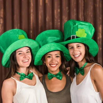 Free image featuring joyful young women wearing St Patrick's Day outfits, including festive hats, and hugging near a wall."