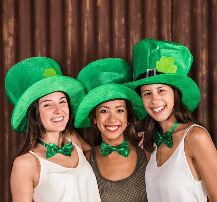 Free image featuring joyful young women wearing St Patrick's Day outfits, including festive hats, and hugging near a wall."