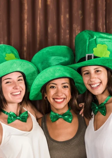 Free image featuring joyful young women wearing St Patrick's Day outfits, including festive hats, and hugging near a wall."