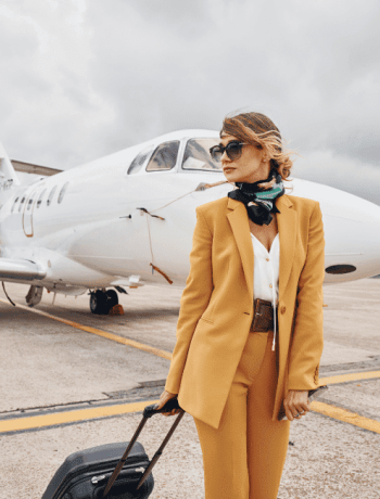 An image of a young woman dressed in stylish yellow airplane outfits stands outdoors near an airplane, holding her luggage