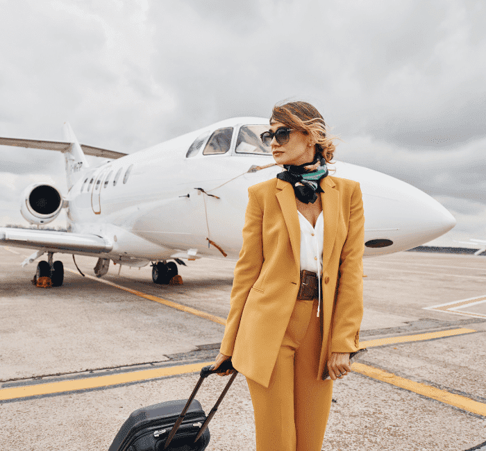 An image of a young woman dressed in stylish yellow airplane outfits stands outdoors near an airplane, holding her luggage