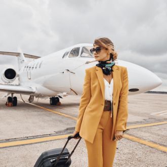 An image of a young woman dressed in stylish yellow airplane outfits stands outdoors near an airplane, holding her luggage