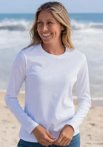 A woman in a white long-sleeve shirt smiles while standing on a sandy beach with ocean waves in the background.