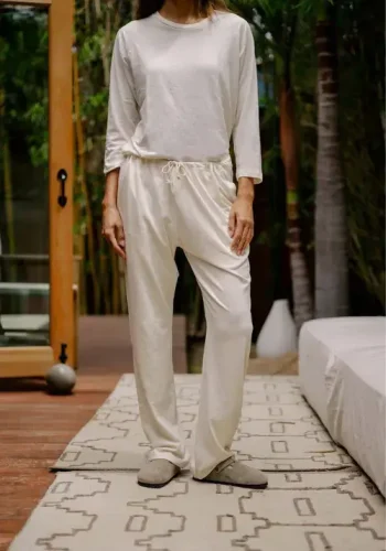 A person wearing a comfortable cream-colored long-sleeve top and matching pants stands indoors on a patterned rug, with a partially open door and greenery in the background.