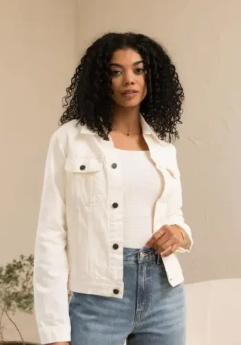 A woman with curly hair wearing a white denim jacket over a white top and blue jeans stands confidently indoors against a neutral background.